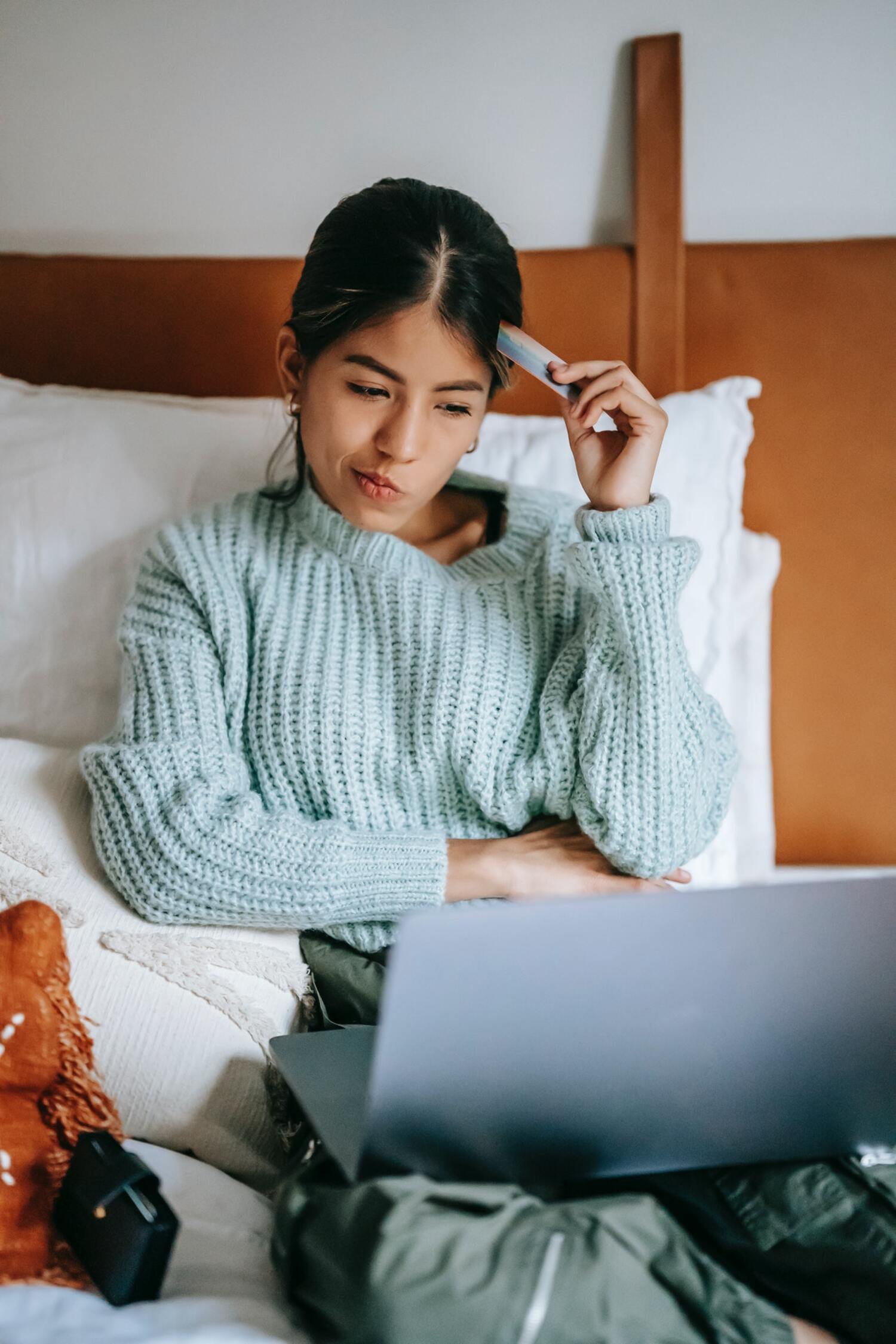 Girl sitting on bed with laptop and credit card thinking