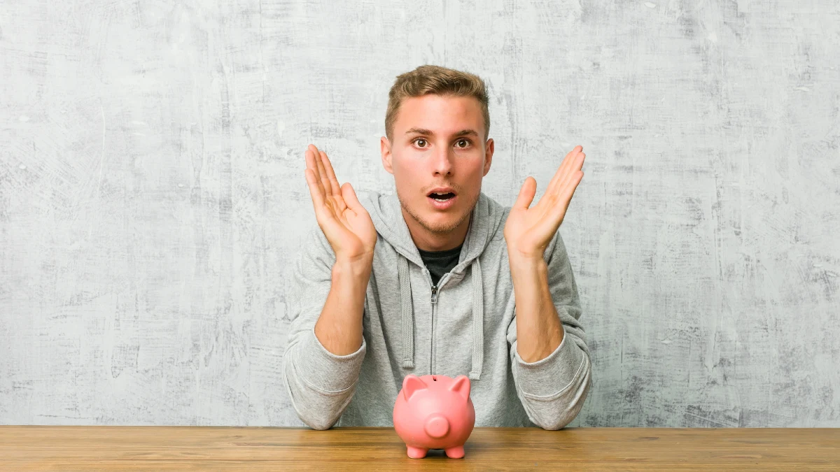 Young man saving money with a piggy bank surprised and shocked